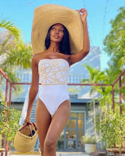Woman wearing a Grand Belmont sun hat, styled in a white swimsuit, walking among tropical plants.