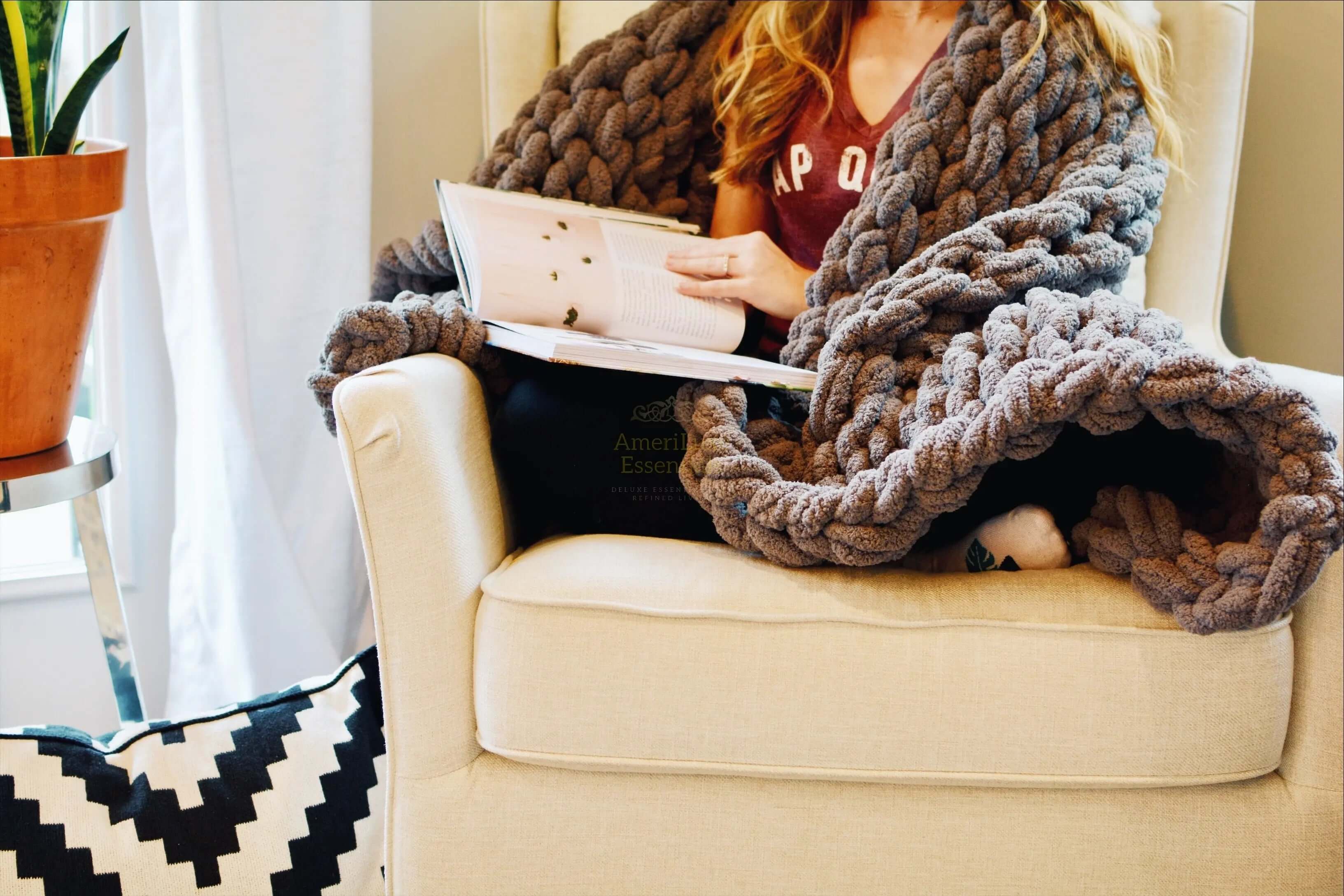 A person cozying up with a dark gray chunky knit blanket, reading a magazine while seated in an armchair.